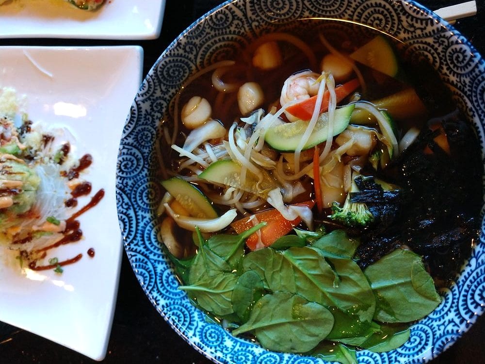 Noodle soup with shrimp, scallops, zucchini, bean sprouts, spinach and nori in a blue-and-white bowl, beside a plate of sauced sushi