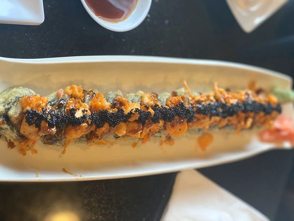 A long tempura-fried roll topped with spicy crab, black tobiko and eel sauce on a narrow white plate