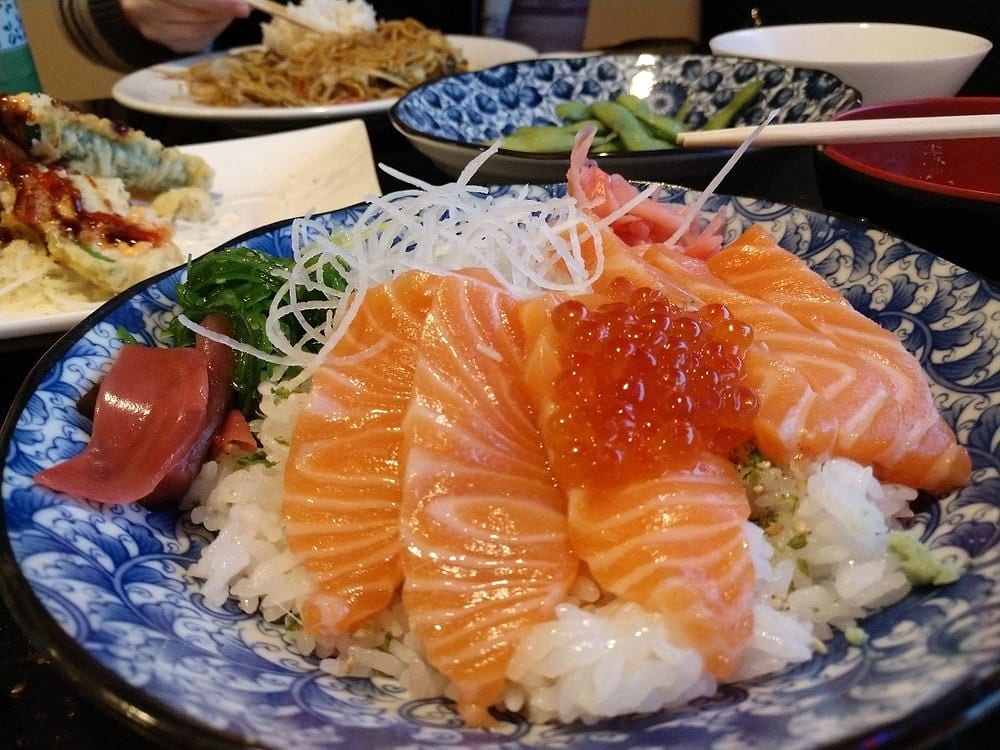 A salmon bowl with sliced salmon, salmon roe, tuna, seaweed salad and daikon over rice, with yakisoba and edamame behind
