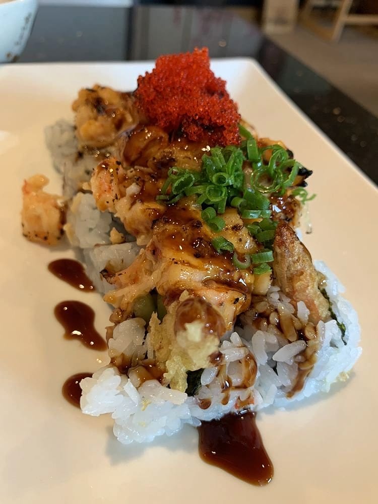 A sushi roll piled with seared shrimp, green onion and red tobiko, with dots of eel sauce on the plate